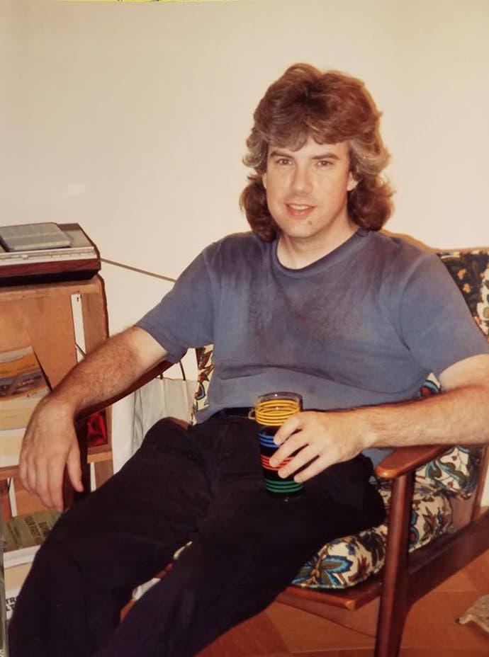 Ein Foto von einem buschigen, behaarten Mann, der in einem gepolsterten Holzstuhl sitzt, eine Tasse mit einem Getränk in der Hand und in die Kamera schaut. Es sieht so verblasst aus, dass es ein Bild aus den 80er Jahren sein könnte, aber es ist aus der Mitte der 90er Jahre. Es ist Lawrence Schick.